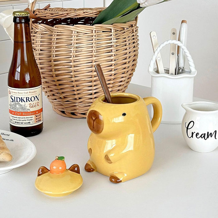 Cute 3D capybara-shaped ceramic coffee mug with lid and small orange detail.
